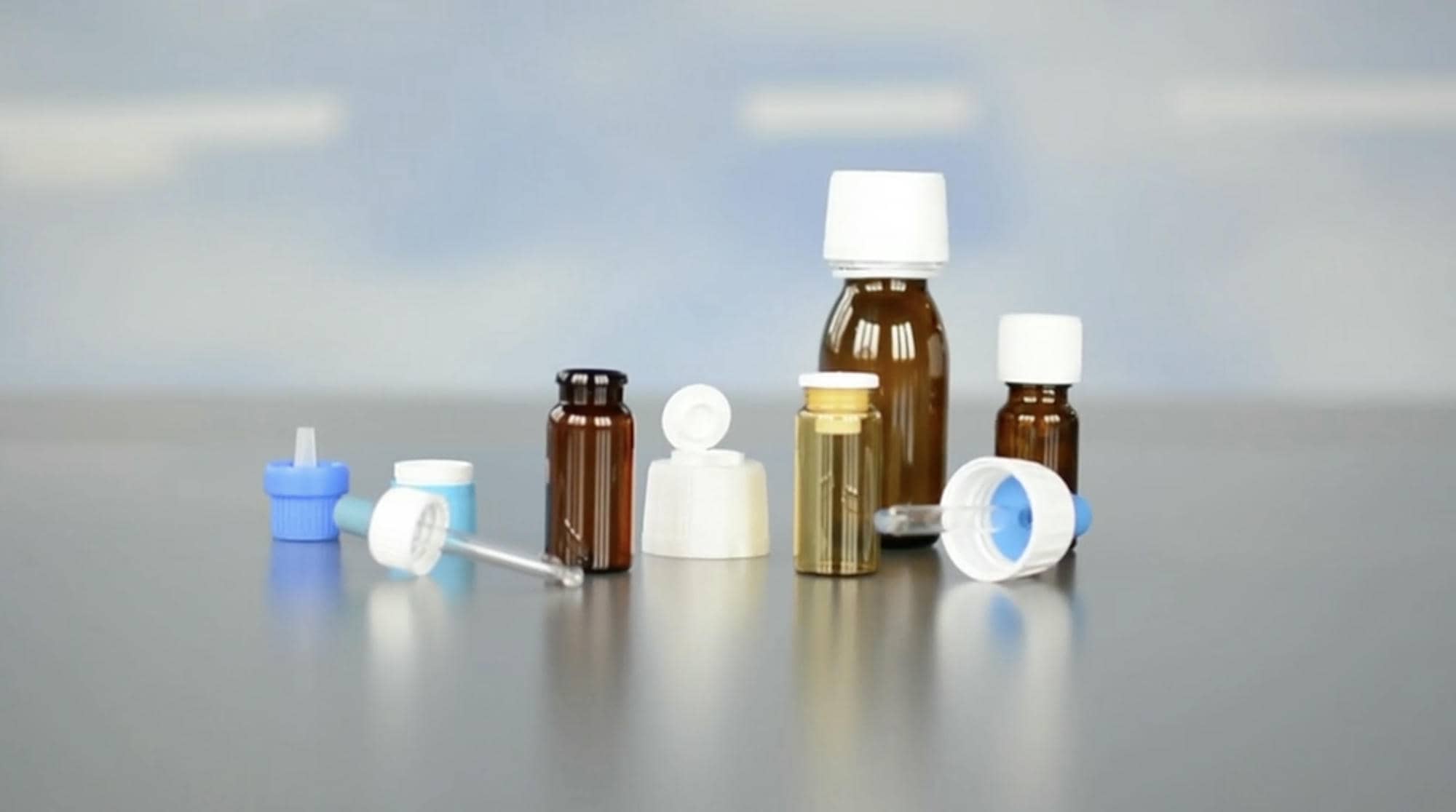 Mehrere pharmazeutische Behälter und Verschlüsse auf einer Oberfläche, darunter braune Glasflaschen, Kunststoffkappen und Dosieraufsätze in Weiß und Blau.