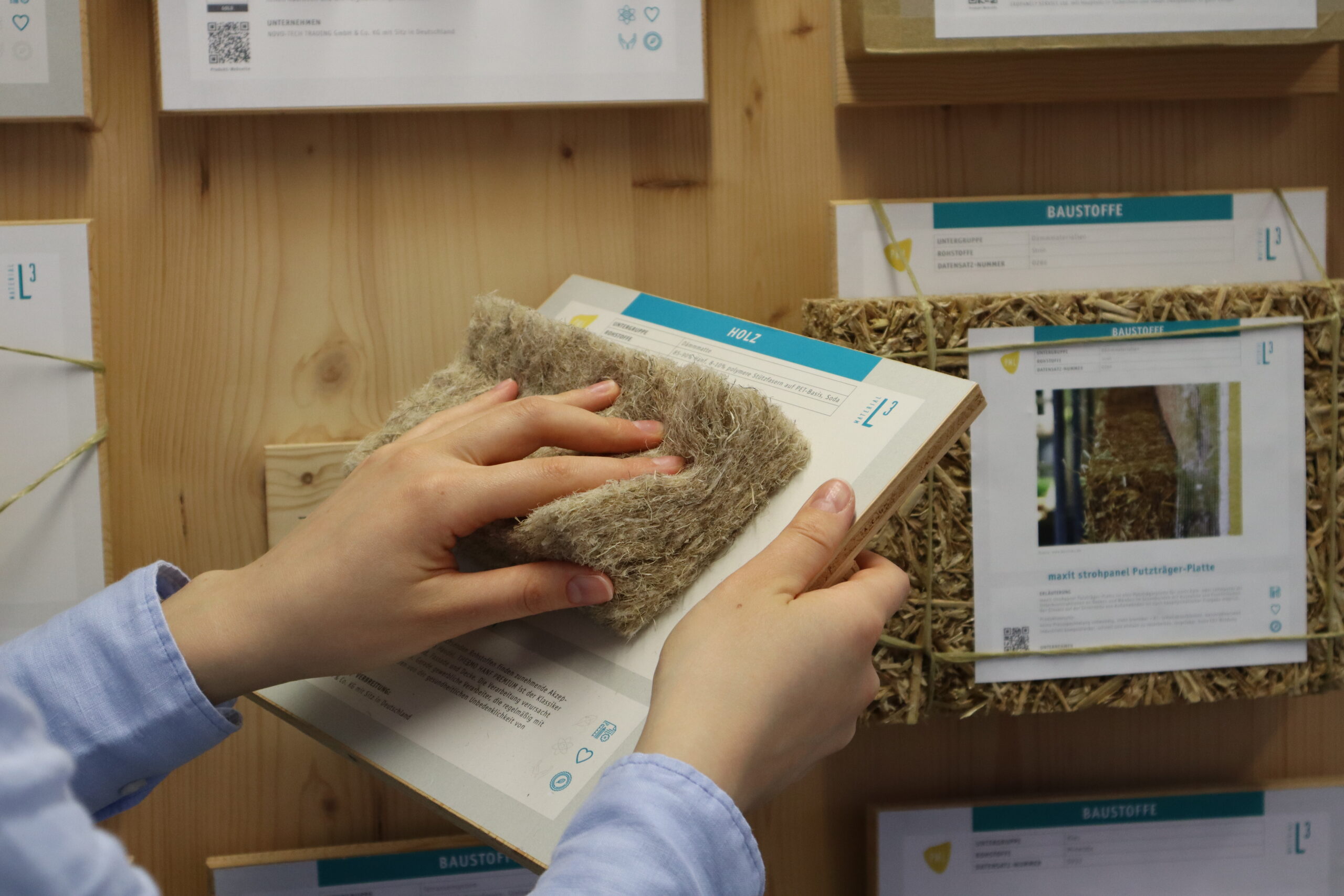 Person hält eine Materialkarte mit Naturfaserdämmstoff vor einer Wand mit dokumentierten nachhaltigen Baustoffproben im Circular Material Lab.